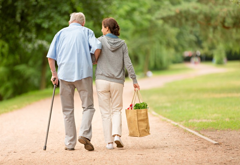 An elderly man with a cane and his adult daughter walk on a path in Potomac MD